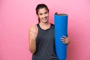 Young Brazilian sport woman going to yoga classes while holding a mat isolated on pink background inviting to come with hand. Happy that you came